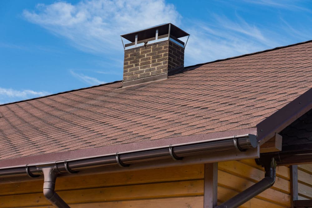 Chimney on a sloped roof against a blue sky with clouds. Roofing shingles visible.